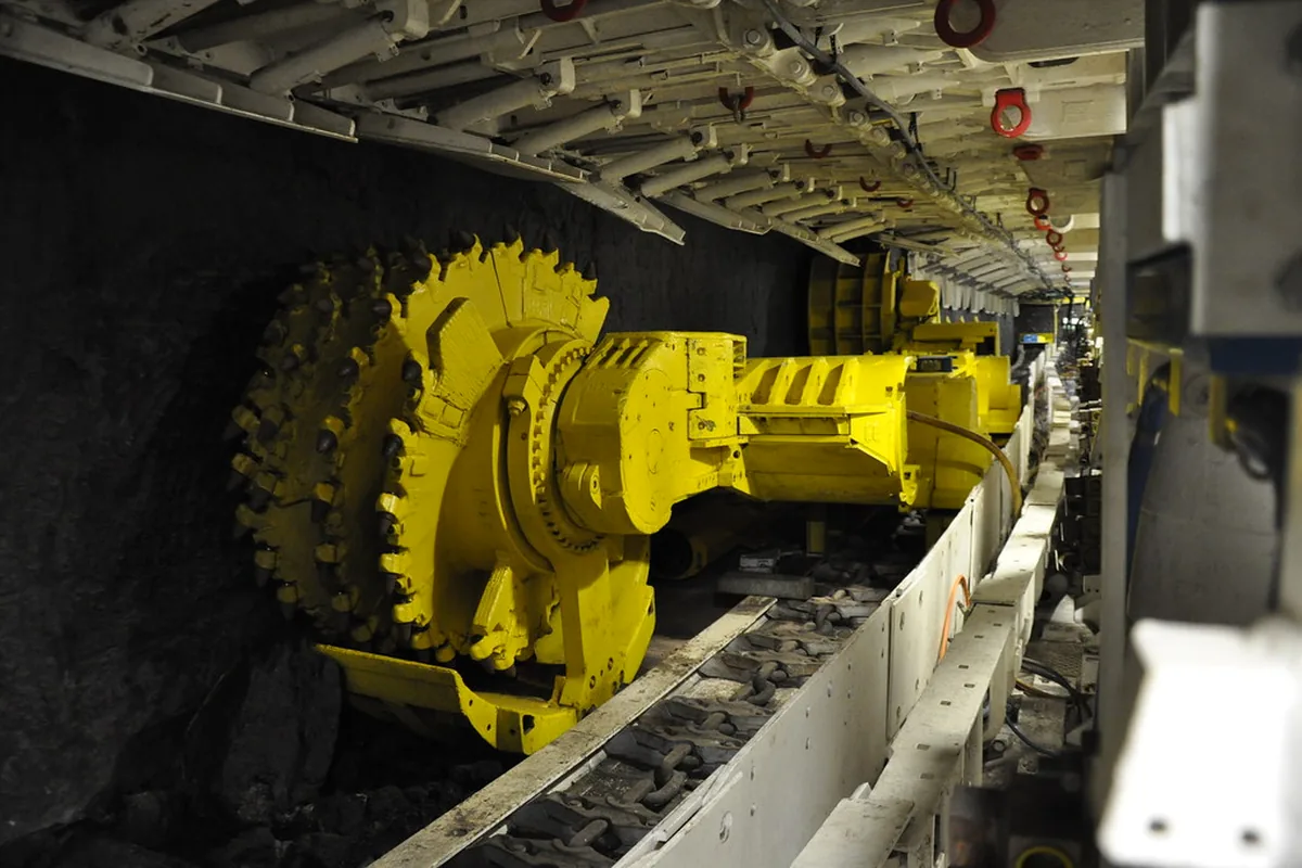 Yellow longwall shearer drum under a row of hydraulic roof supports beside the face conveyor in a coal mine, the kind of panel whose gate-road equipment MGM 109 Type 1013 cables supply
