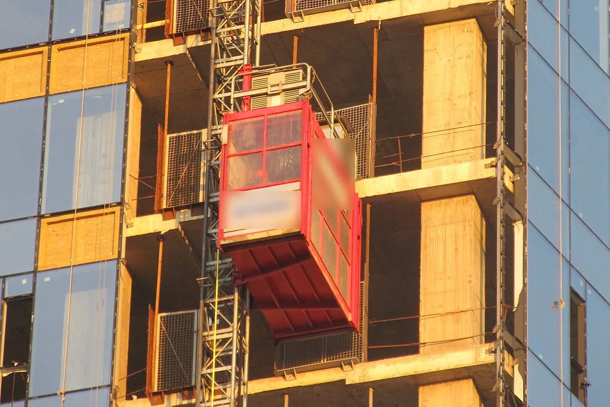 Rack-and-pinion construction hoist car on a mast climbing the face of a high-rise under construction, the kind of lift fed by a BAULIFT power and control cable