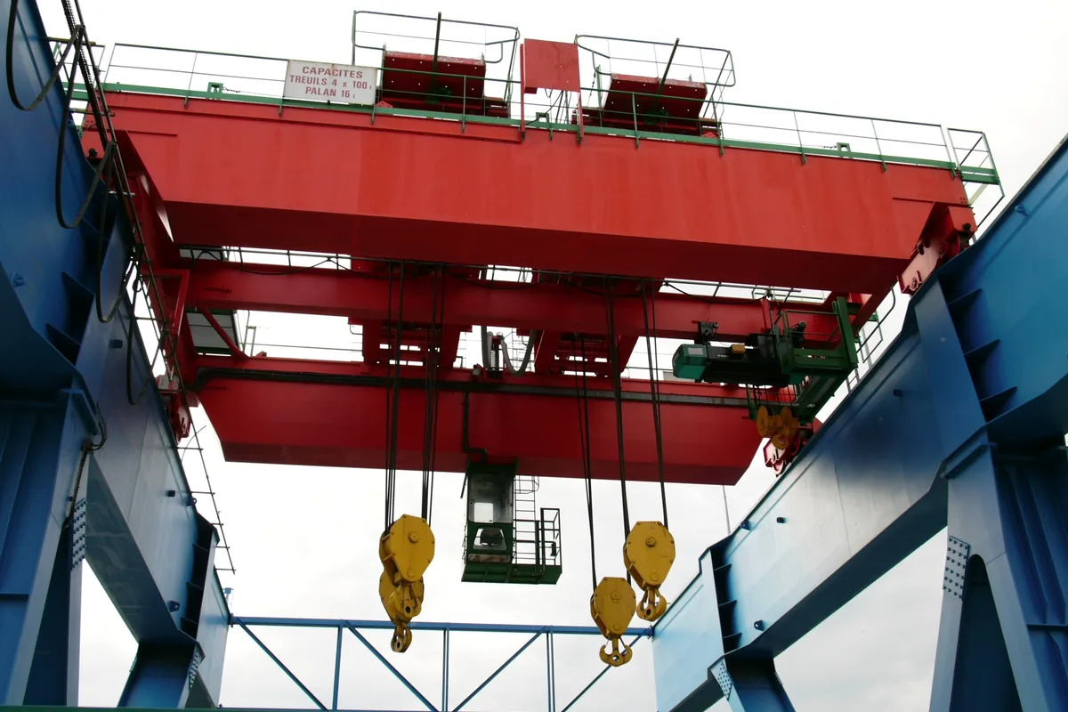 Red double-girder overhead crane between blue gantry legs, with flat cables hanging in festoon loops from trolleys at the top left, the duty an NGFLCGÖU-J festoon cable is built for