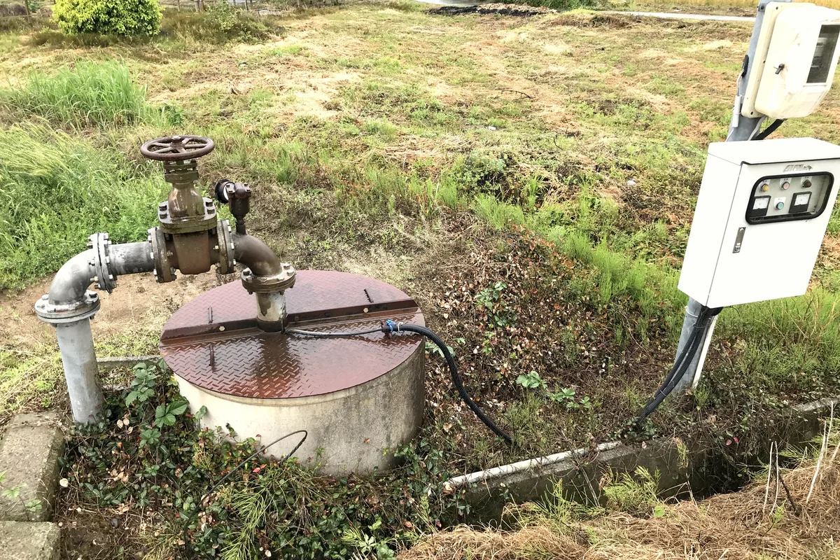 Electric water well beside farmland, with the well head, a discharge valve and a black cable running from the well to the pump control panel, the route an S1BNH2-F cable takes