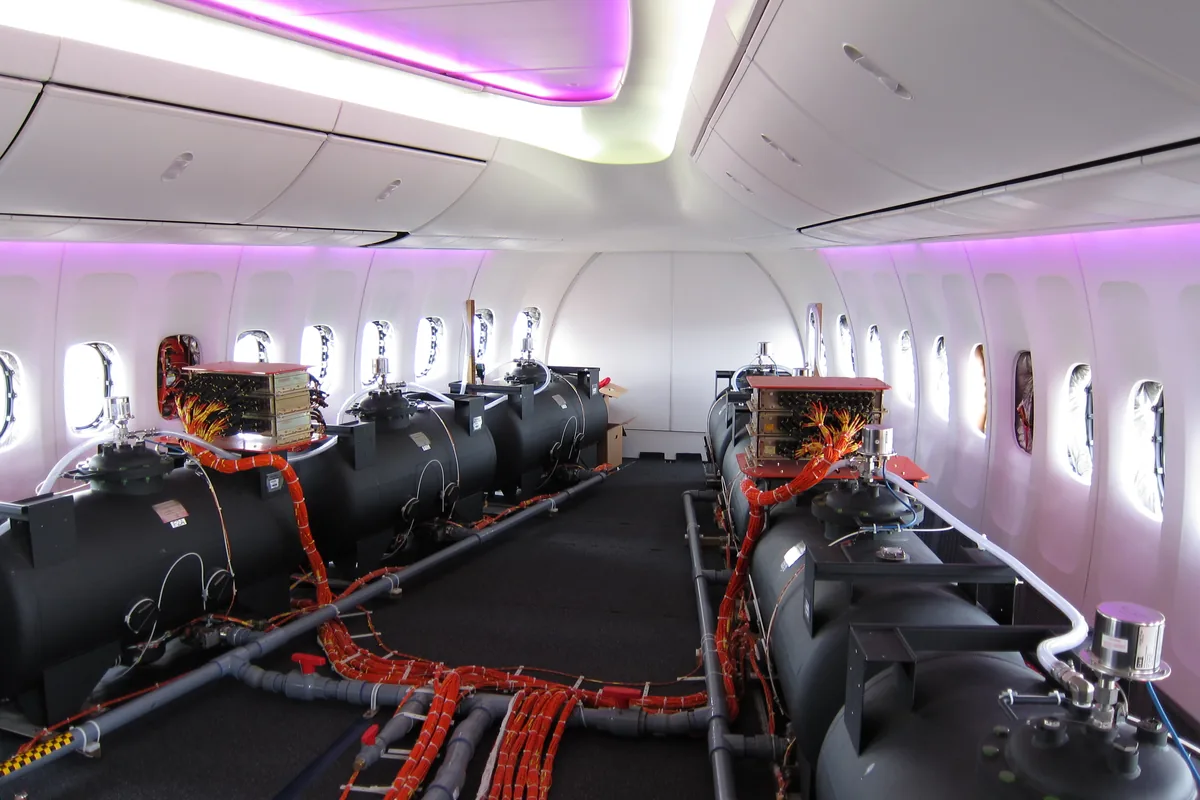 Flight-test instrumentation in an airliner cabin: data-acquisition units on top of ballast tanks, with bundles of orange sensor wiring routed along the floor